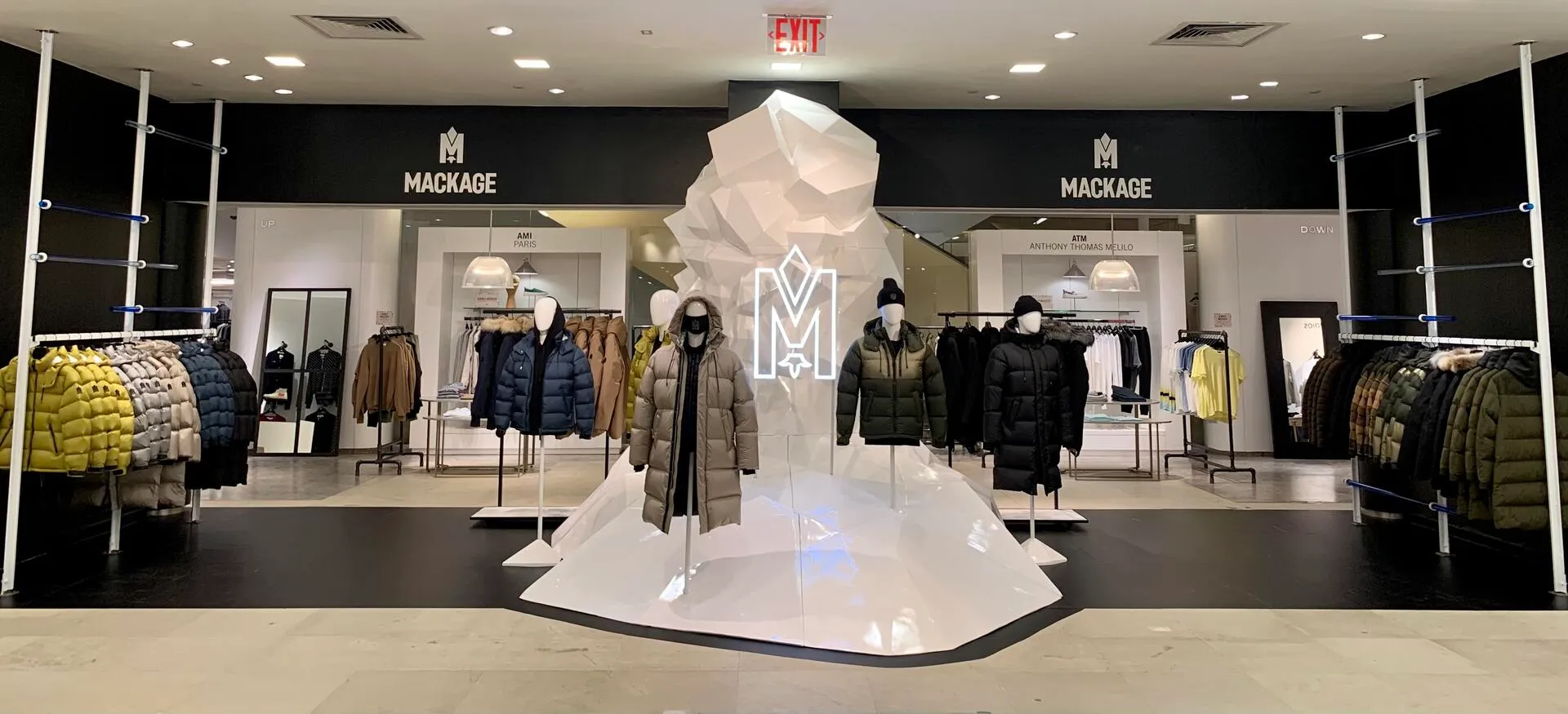 Mackage men's pop-up at Saks Fifth Avenue: a faceted white avalanche sculpture with an illuminated M logo, surrounded by mannequins in down parkas.
