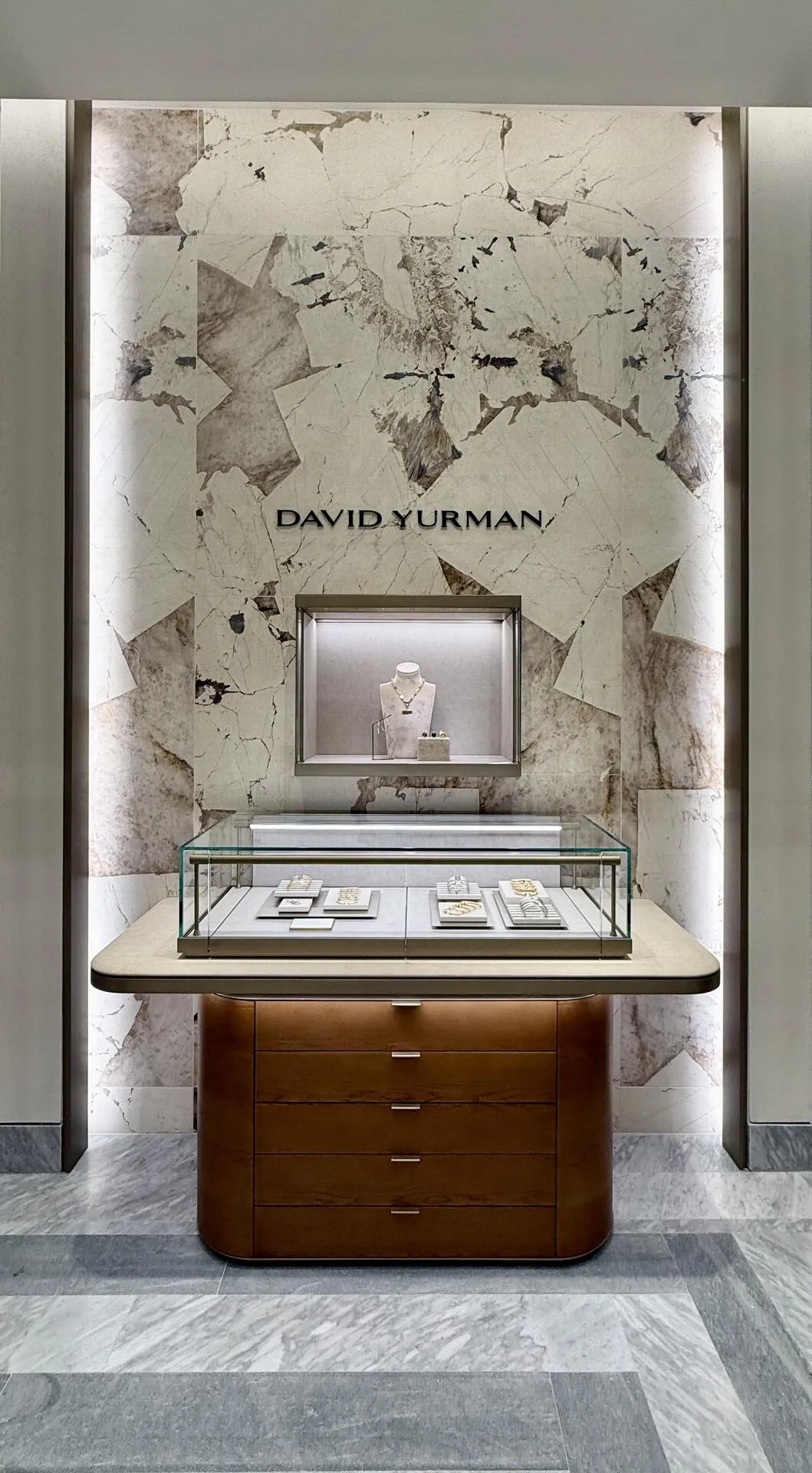 David Yurman feature wall in book-matched stone with a lit niche holding a necklace, above a glass case of Cable bracelets on a walnut cabinet.