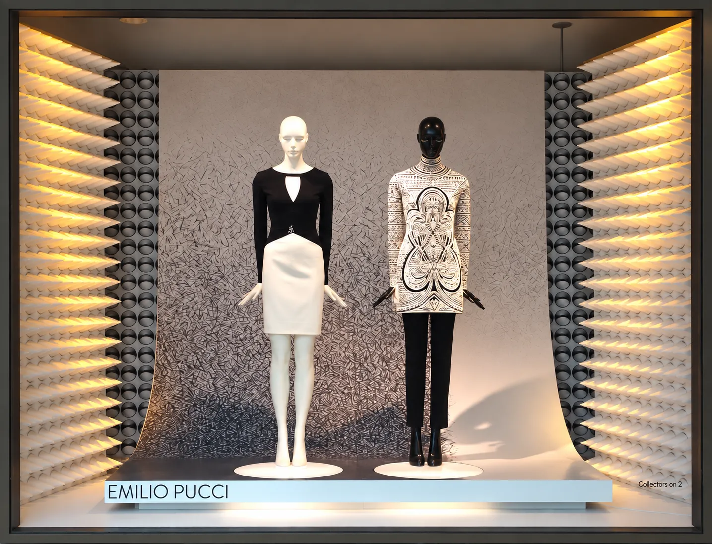 Emilio Pucci window: two mannequins, one in a black-and-white dress and one in a graphic-print tunic with black trousers, stand before a hand-drawn black-and-white backdrop between glowing sculptural fin walls.