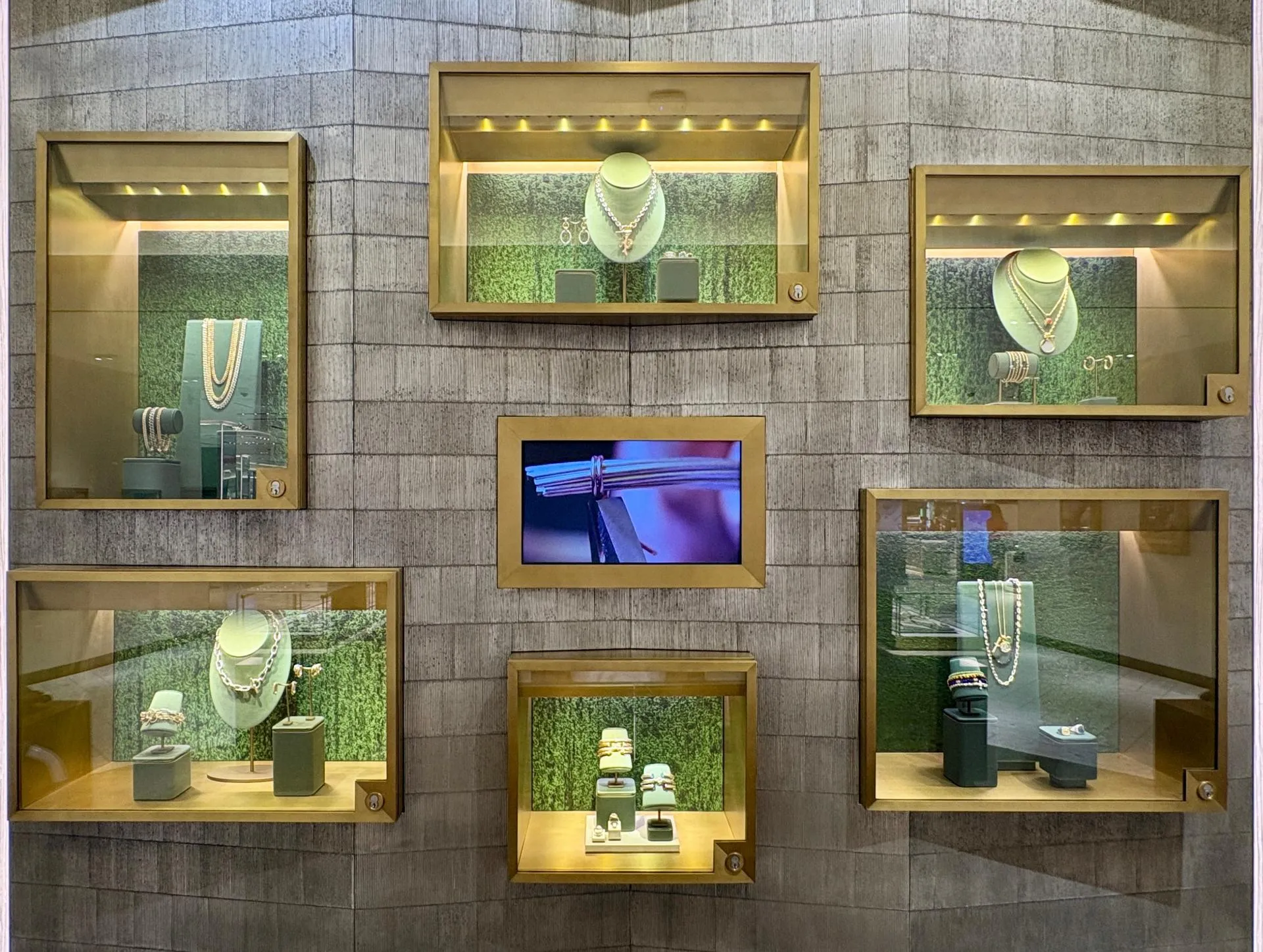 A wall of brass-framed jewelry vitrines with moss-green backdrops, arranged around a central screen showing a Cable bracelet being made.