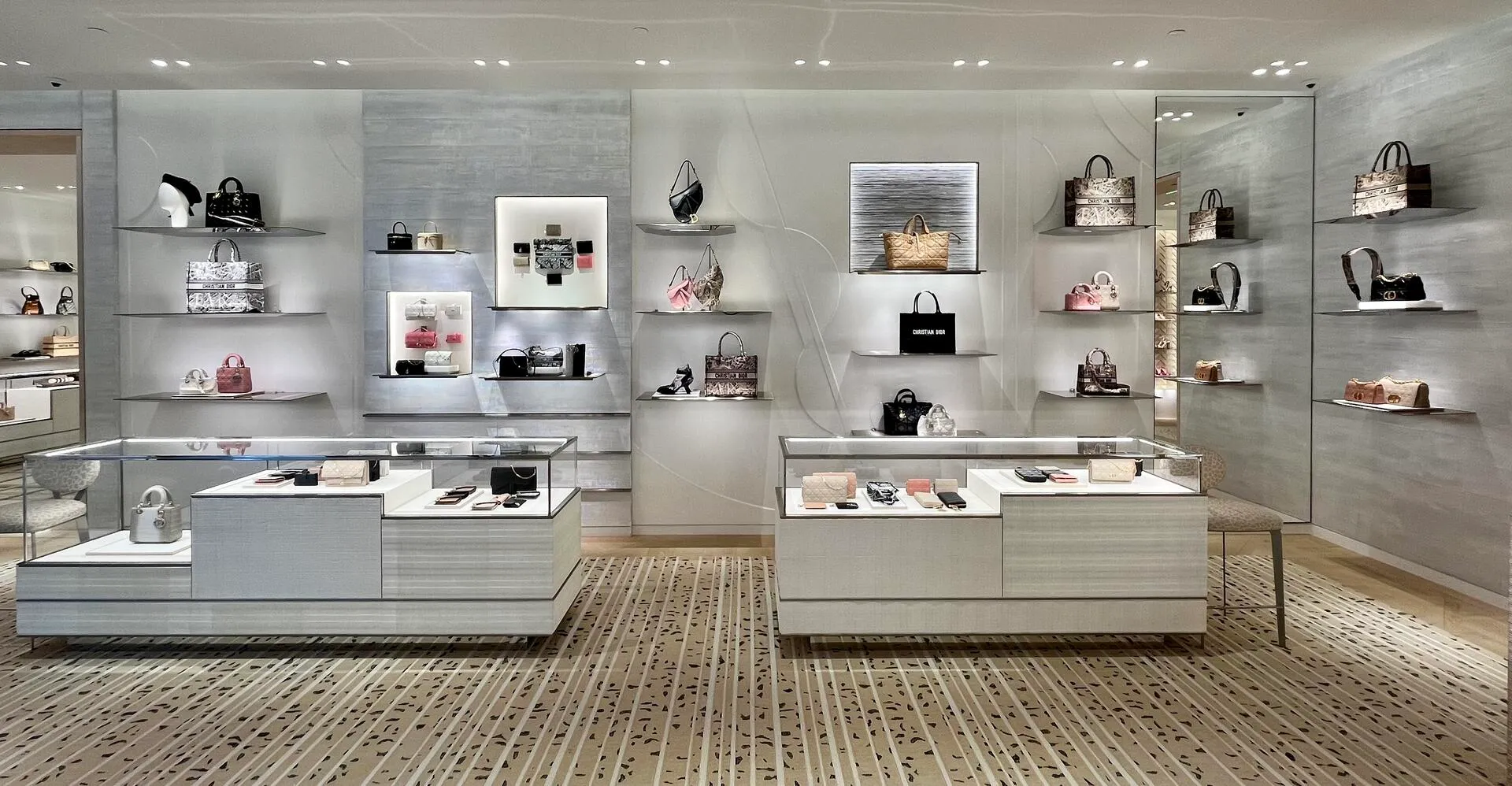Dior leather goods salon with handbags presented on floating shelves and in illuminated niches above two white display cases.