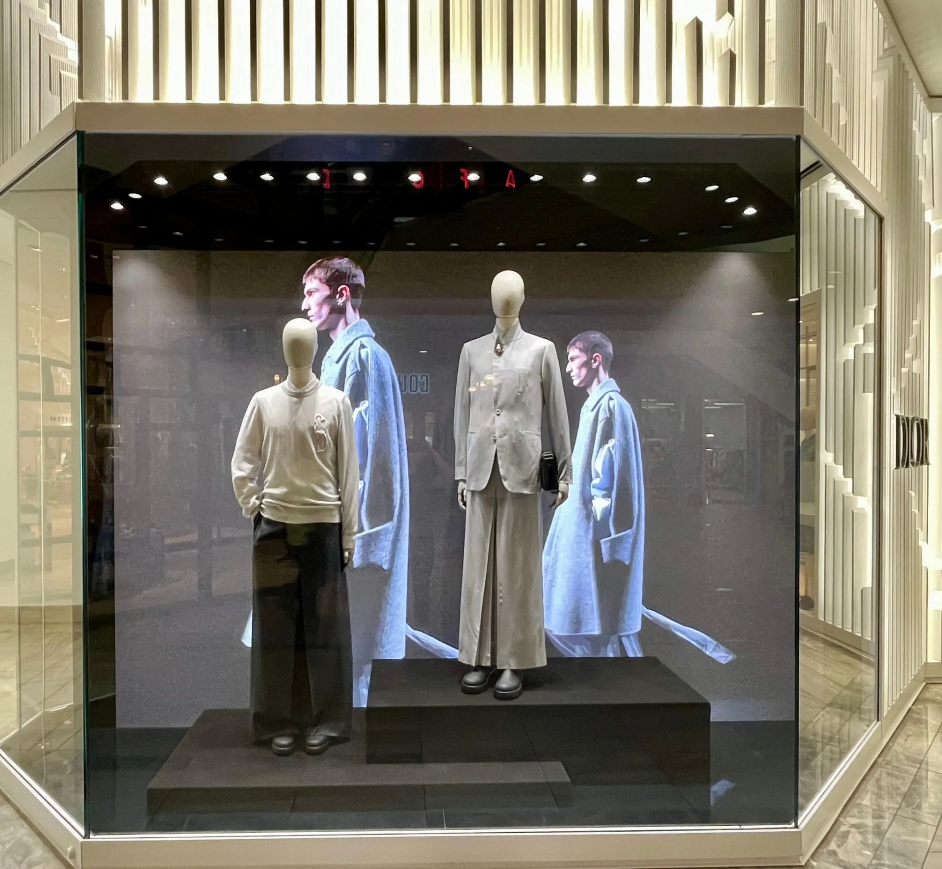 Dior men's window with two mannequins in tonal tailoring on black plinths, set against large-scale campaign imagery.