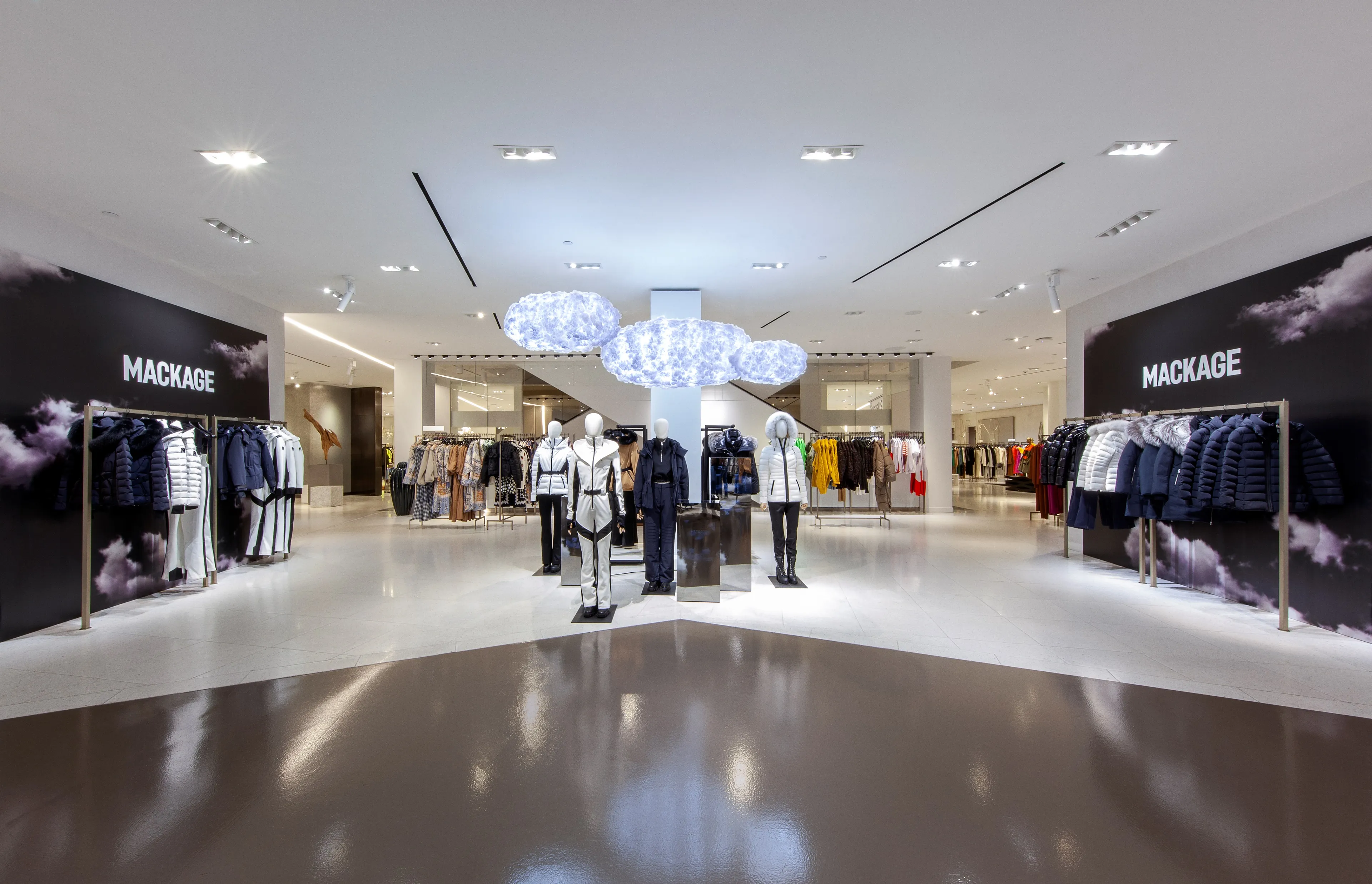 Mackage cloud pop-up in the fifth-floor women's atrium at Saks Fifth Avenue New York: midnight-sky cloud walls framing mannequins in winter outerwear beneath illuminated cloud forms.