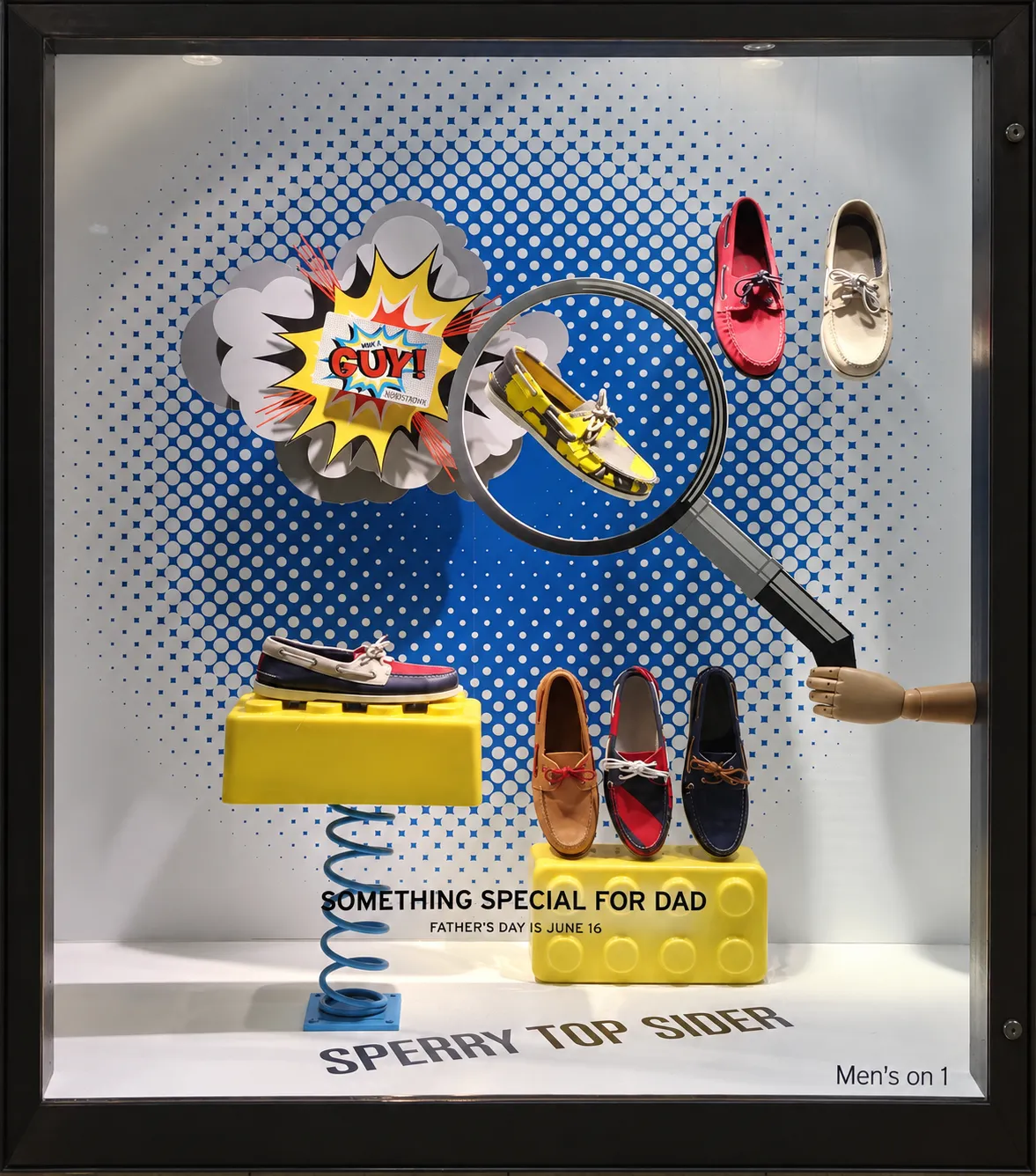 Sperry Top-Sider Father's Day window: boat shoes on oversized yellow toy bricks and a coiled spring against a blue pop-art halftone backdrop, with a comic-book burst and a giant magnifying glass held by a wooden hand.