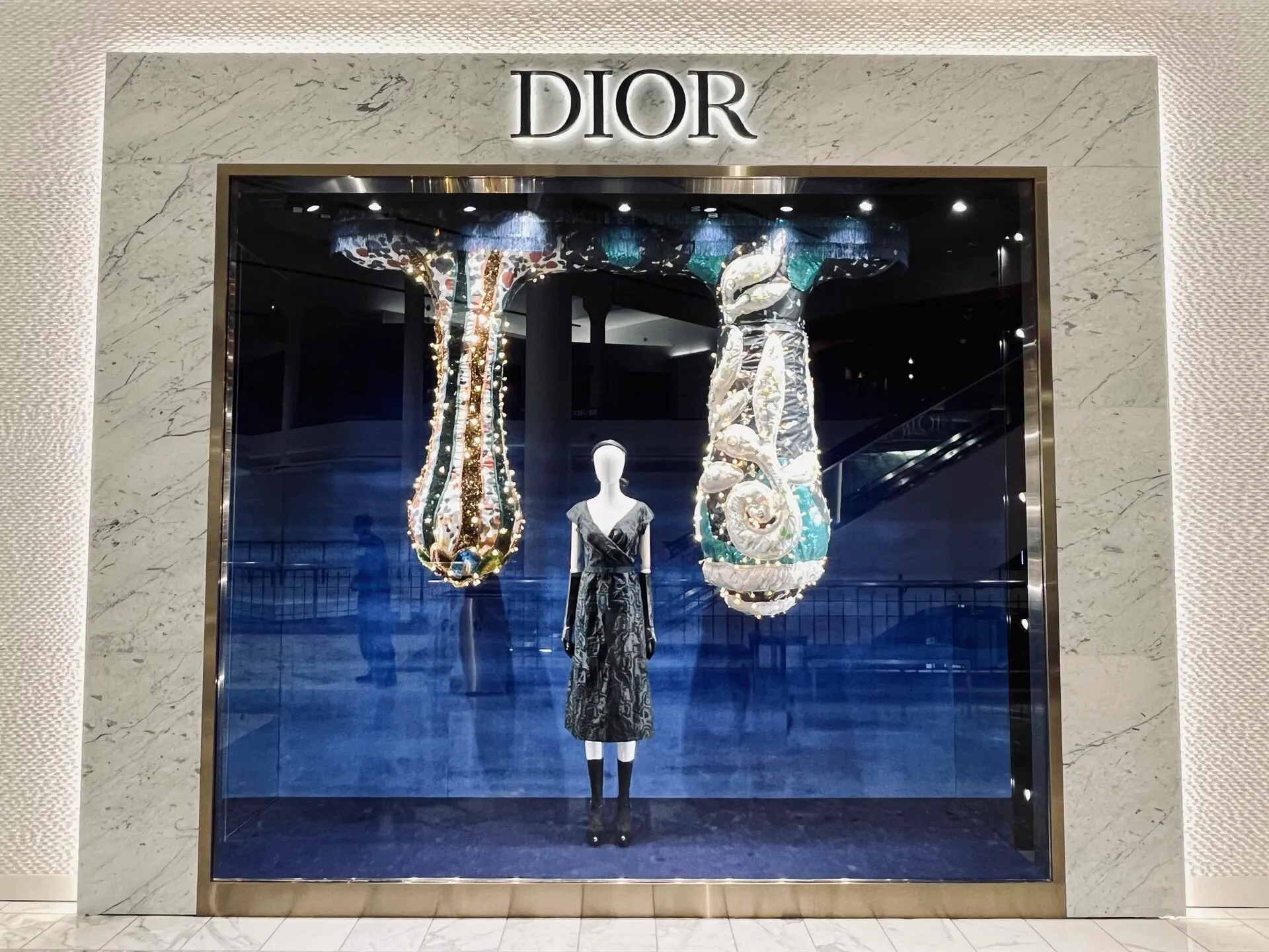 Dior boutique window: a mannequin in a black dress between two illuminated, sculptural hanging installations against a deep blue backdrop.